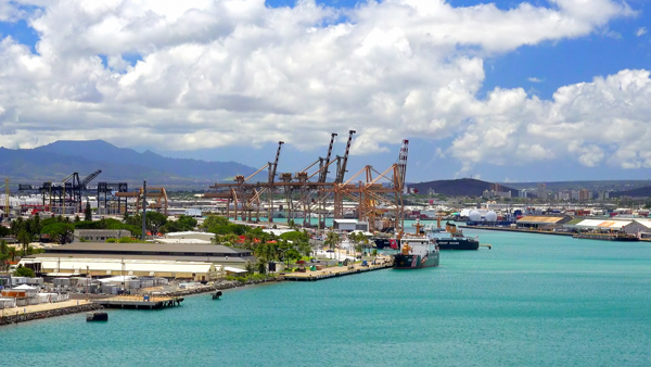 View of a bustling shipping port in Hawaii with cargo ships and cranes against a backdrop of blue skies and mountains.